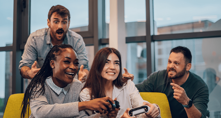 Designing Digitally - Four people are sitting on a couch, two of them playing video games while the other two cheer them on. They appear to be enjoying themselves in a casual indoor setting, much like employees might during a relaxed team-building session with an engaging elearning vendor. Designing Digitally -