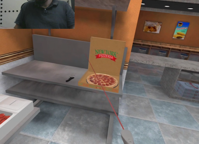Designing Digitally - A virtual reality view of a person pointing a laser towards an open pizza box labeled "New York Pizzeria," containing a partially eaten pizza, all situated on a counter in a simulated kitchen designed for fast food training simulations. Designing Digitally -