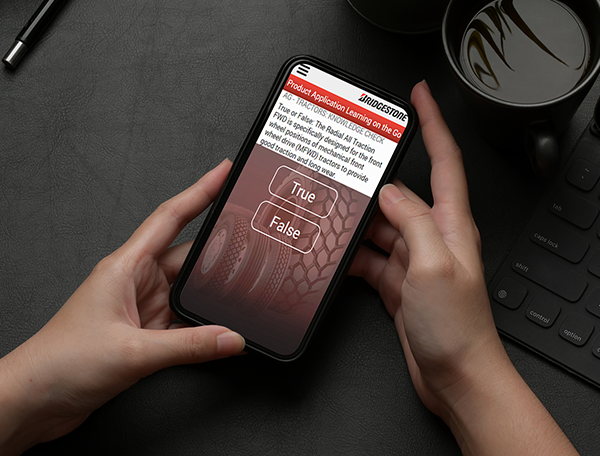 A person holding a smartphone with a true/false quiz on the Mobile Application Training screen, placed on a desk next to a keyboard and a cup of coffee.
