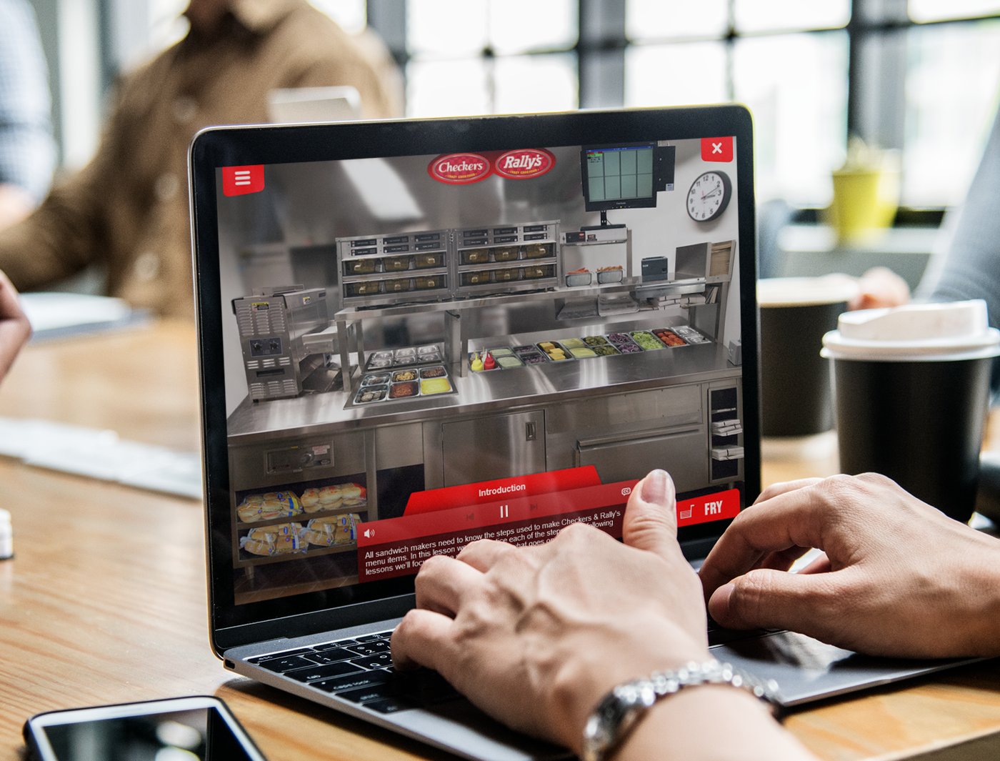 Laptop screen displaying a virtual fast-food ordering interface for Custom Training Games for Employees, with user's hands interacting with the website.