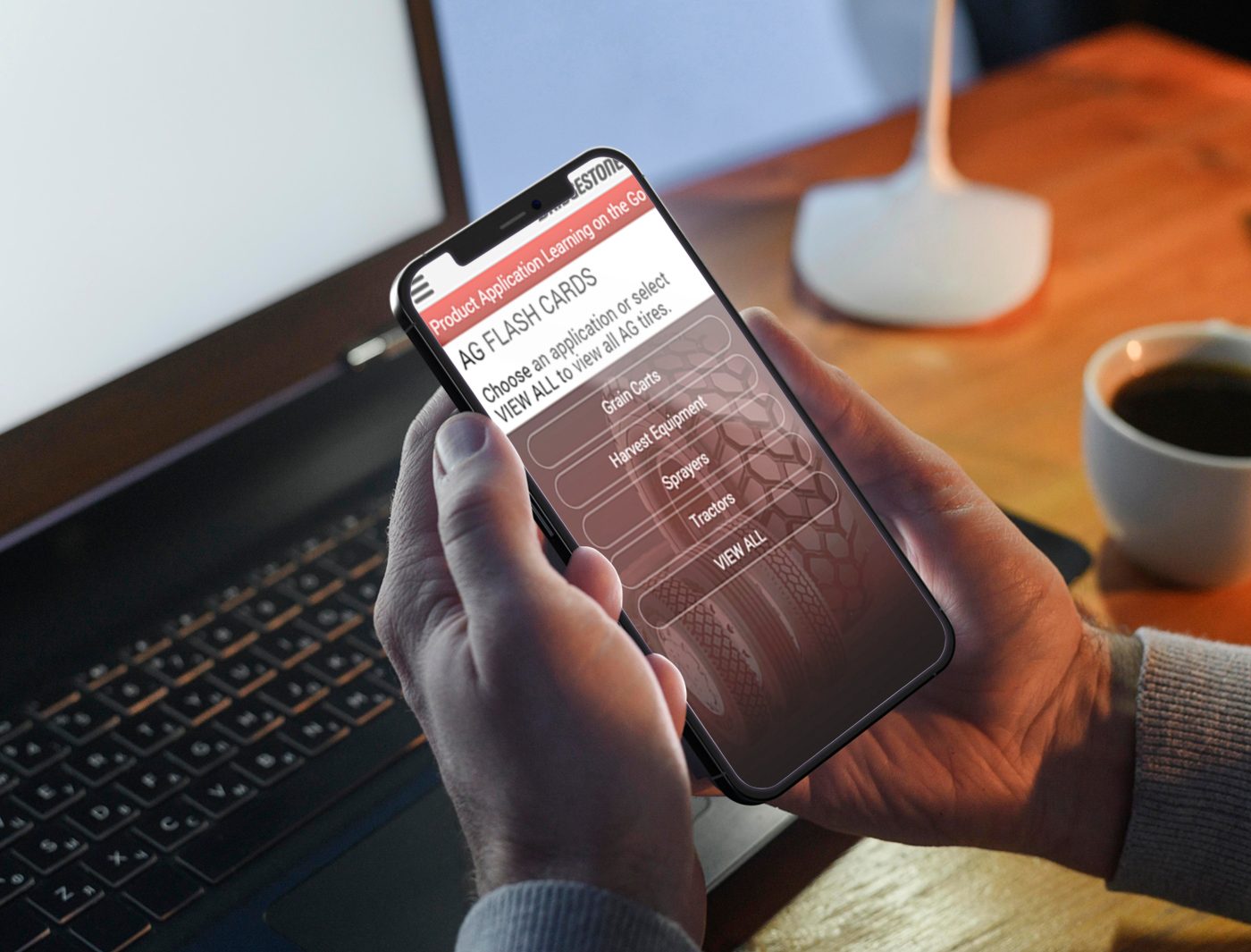 A person using a mobile product application on a smartphone for training with a laptop and a cup of coffee in the background.