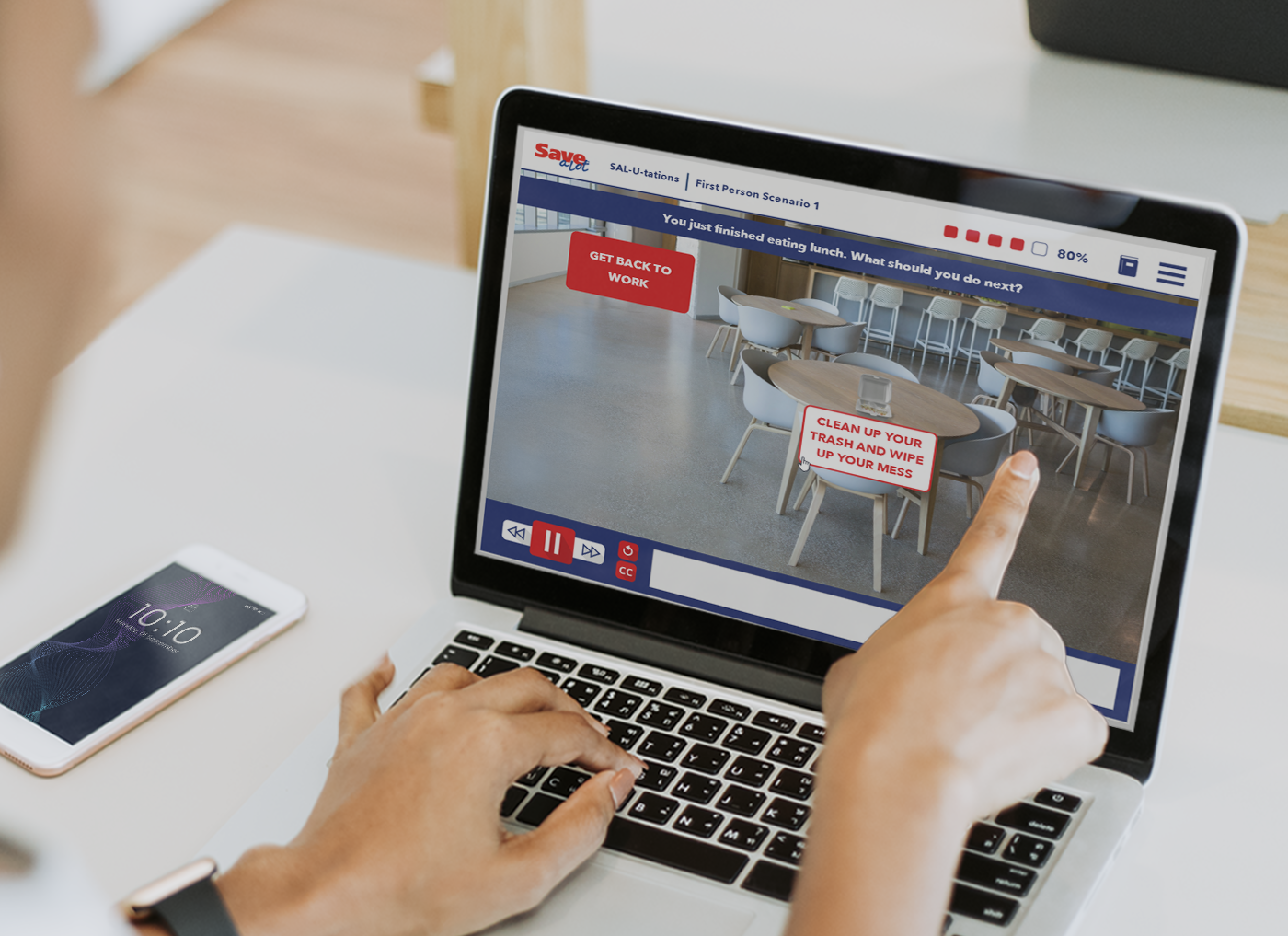 Designing Digitally - A person's hands using a laptop showing a virtual reality classroom scene with interactive buttons saying "get to work" and "clean up your trash and wipe your hands". Designing Digitally -