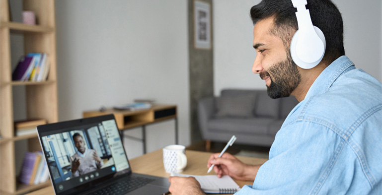 Designing Digitally - A man wearing headphones participates in an online training session on a laptop while taking notes, sitting at a desk with a mug next to him. Designing Digitally -