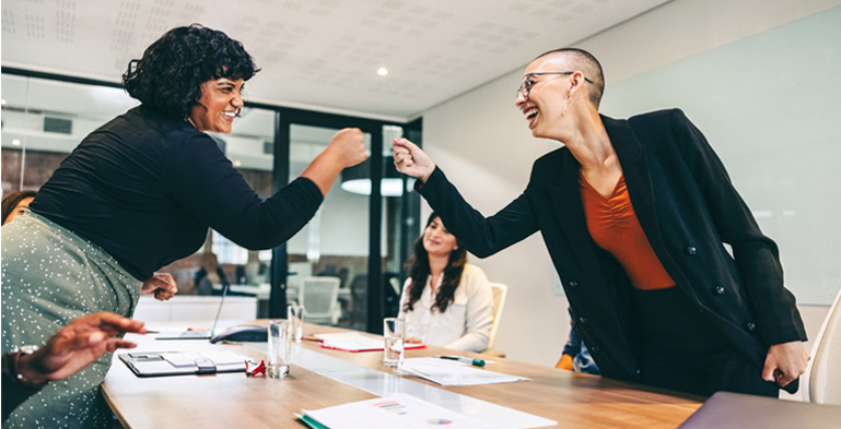 Designing Digitally - Two women fist bumping joyfully during a business meeting while colleagues watch, reflecting a positive atmosphere in a modern office setting with Virtual Reality simulations. Designing Digitally -