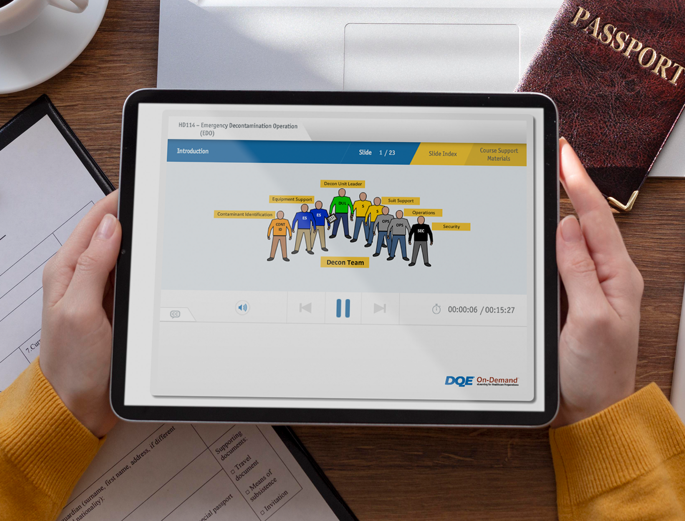 Designing Digitally - Hands holding a tablet displaying an eLearning vendor's corporate training presentation on teamwork, with documents and a passport on the desk. Designing Digitally -