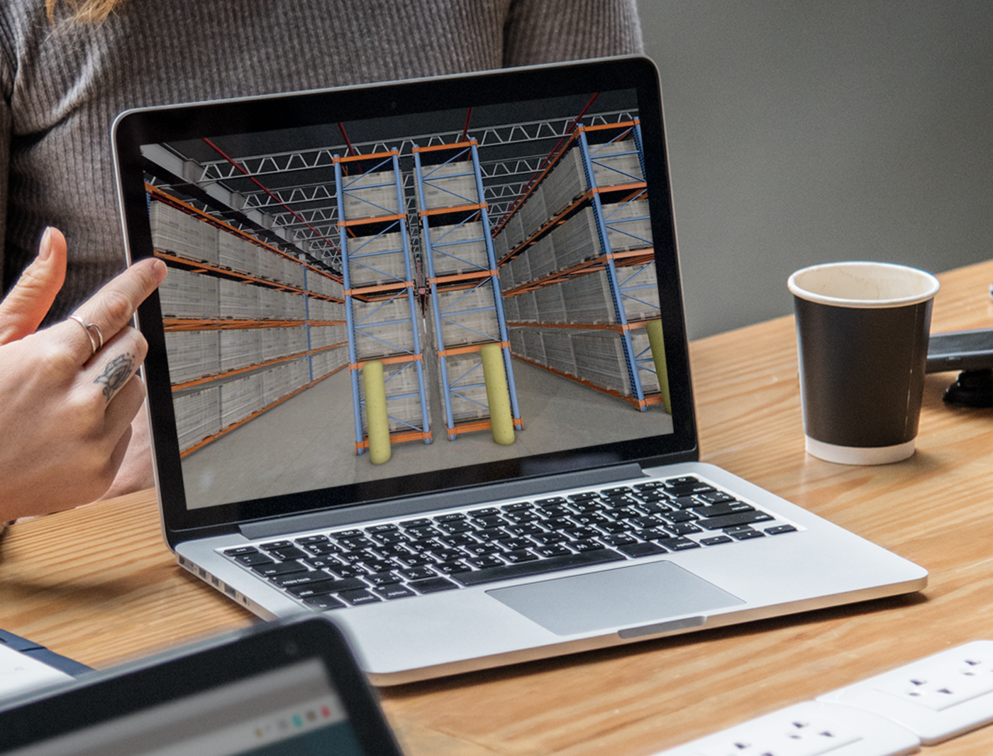 Designing Digitally - A person pointing at a laptop screen displaying an image of a large warehouse with metal shelving. A coffee cup and keyboard are also on the table, alongside icons of a mobile app for warehouse management. Designing Digitally -