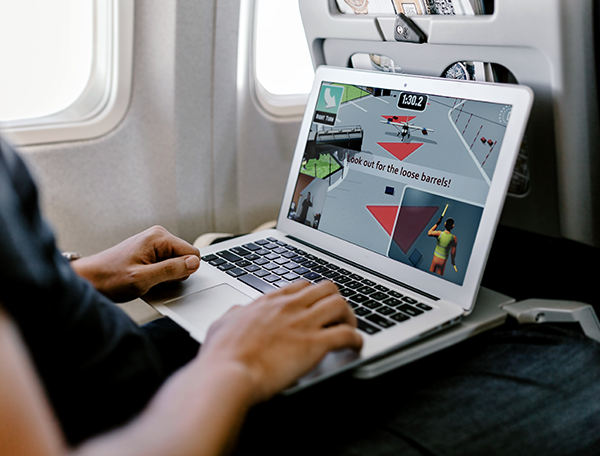 Designing Digitally - A person is playing a computer game on a laptop while sitting in an airplane seat, focused intently as the game shows an alert to watch out for loose barrels. They seem deeply engaged, almost as if they are participating in training games using motion tracking technology. Designing Digitally -