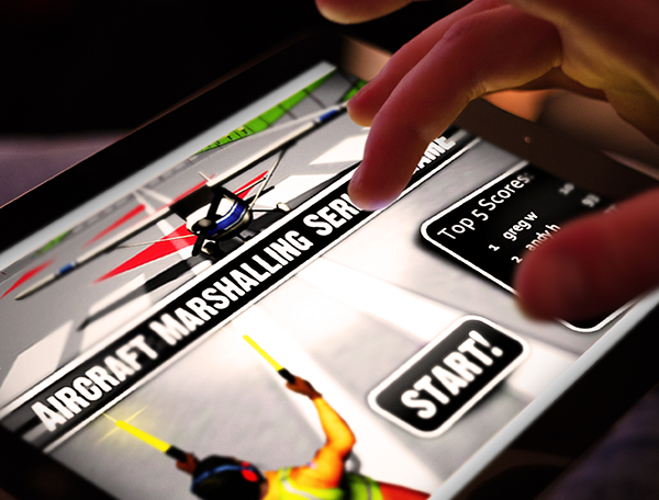 Designing Digitally - Close-up of a person using a touch screen device displaying an aircraft marshalling simulation training game with "Start!" and "Top 5 Scores" visible on the screen, incorporating motion tracking technology for an immersive experience. Designing Digitally -