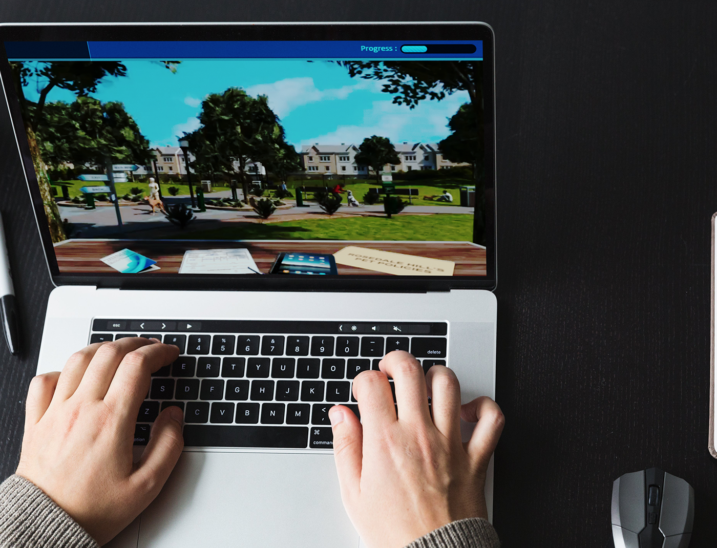 Designing Digitally - Hands typing on a laptop keyboard with a pet ownership training simulation game showing a park scene on the screen; mouse and pen are visible on the dark surface beside the laptop. Designing Digitally -