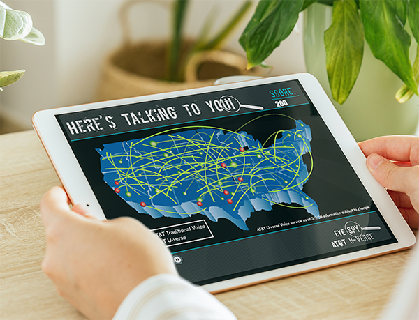 Designing Digitally - Person holding a tablet displaying a map of the United States with interconnected lines and text reading "Here's Talking to You!" and a score of 200, highlighting trauma nurse training simulations. The background shows plants on a table. Designing Digitally -