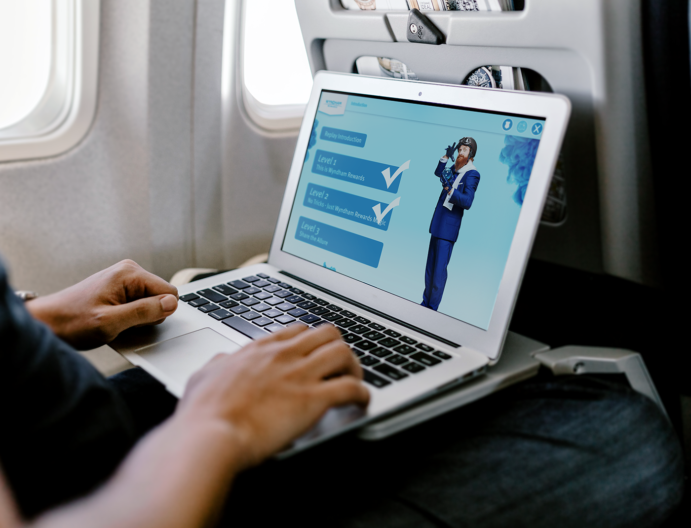 Designing Digitally - A person is sitting in an airplane seat, using a laptop that displays an online course with levels of English instruction, as part of a customer rewards training program for Wyndham Hotels. Designing Digitally -