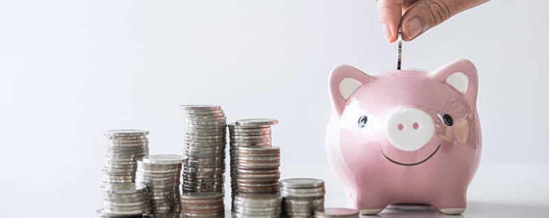 Person saving money by inserting a coin into a piggy bank with stacks of coins beside it.