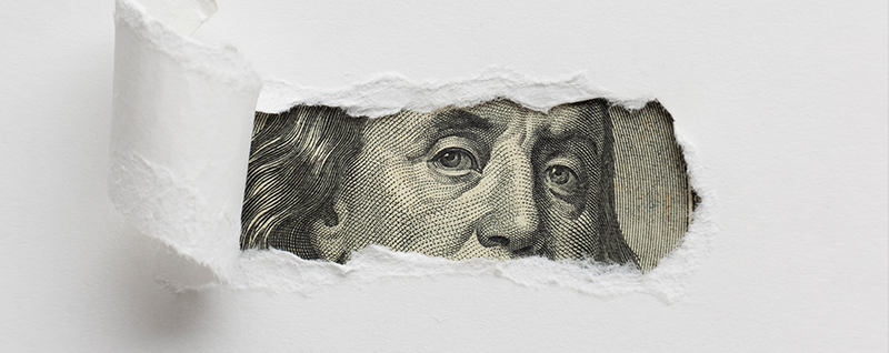 Designing Digitally - A torn piece of white paper reveals a portion of a $100 bill, showing Benjamin Franklin's eyes and part of his face, much like discovering the value behind an effective online training vendor. Designing Digitally -