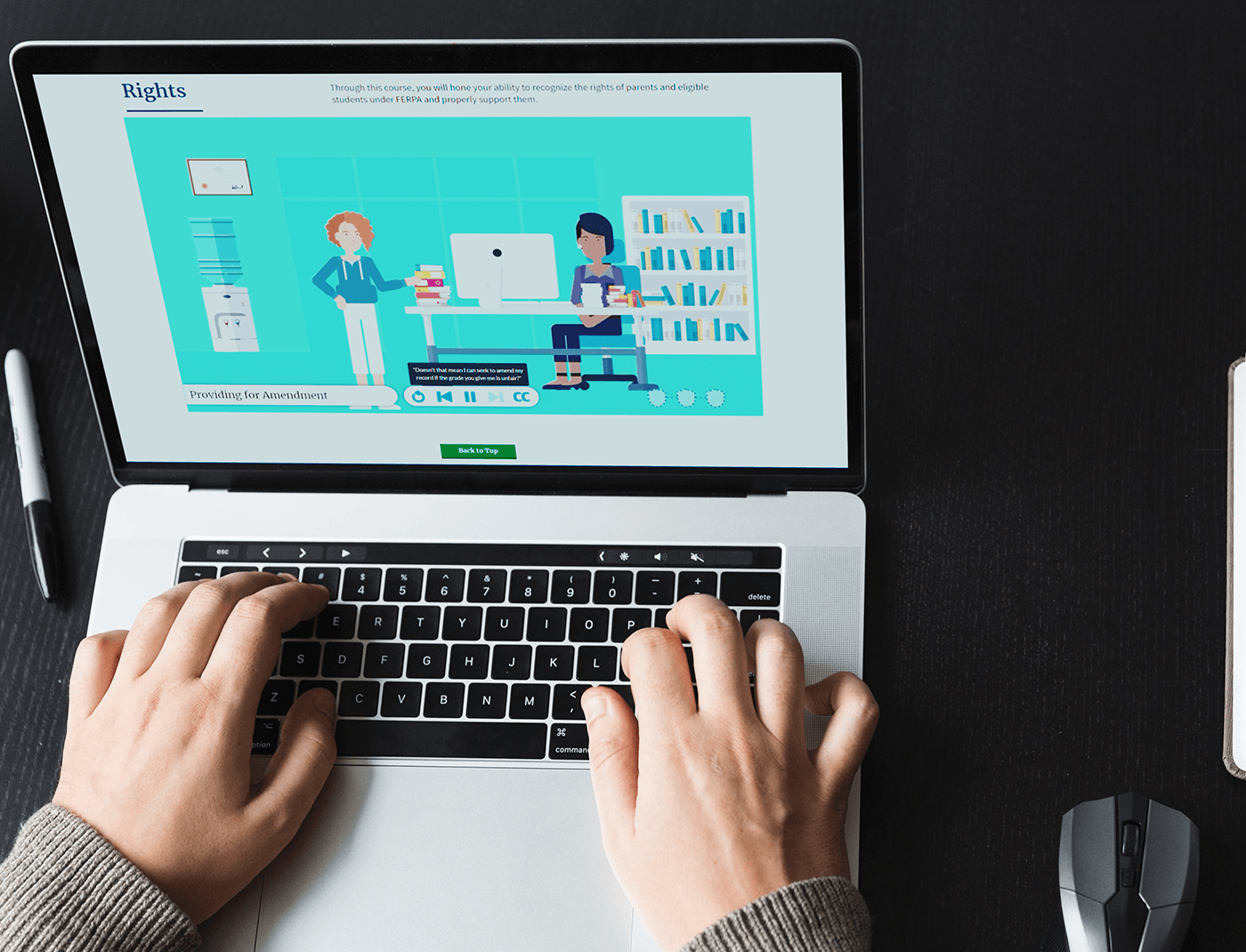 Designing Digitally - Person typing on a laptop, watching an online educational video featuring animated characters in a classroom setting, with a monitor, desk, chair, and bookshelves in the background. The course includes occupational therapy simulations to enhance the learning experience. Designing Digitally -