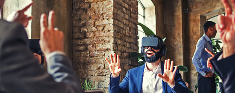 Designing Digitally - A man wearing a virtual reality headset sits with raised hands in an industrial-style office environment, while another man in the background is looking at his phone. Designing Digitally -