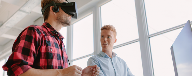 Designing Digitally - Two individuals are in an office. One wears a virtual reality headset, immersed in simulation learning, while the other, standing near a desk, looks on attentively. The background shows large windows with ample natural light, highlighting the innovative approach of this vendor. Designing Digitally -