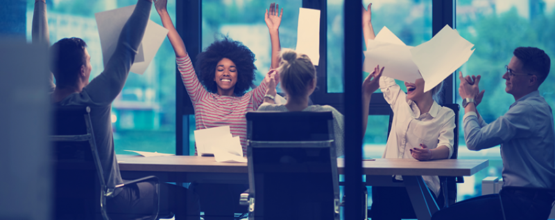 Designing Digitally - A group of six people sitting around a table in an office cheerfully tossing papers into the air, celebrating the successful integration of gamification into their mobile learning platform. Designing Digitally -