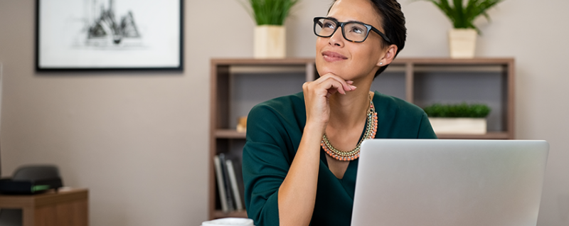 Designing Digitally - A person wearing glasses and a green shirt, seated at a desk with a laptop, looks thoughtfully to the side. There are potted plants and a framed picture in the background, creating an ideal environment for workplace learning. Designing Digitally -